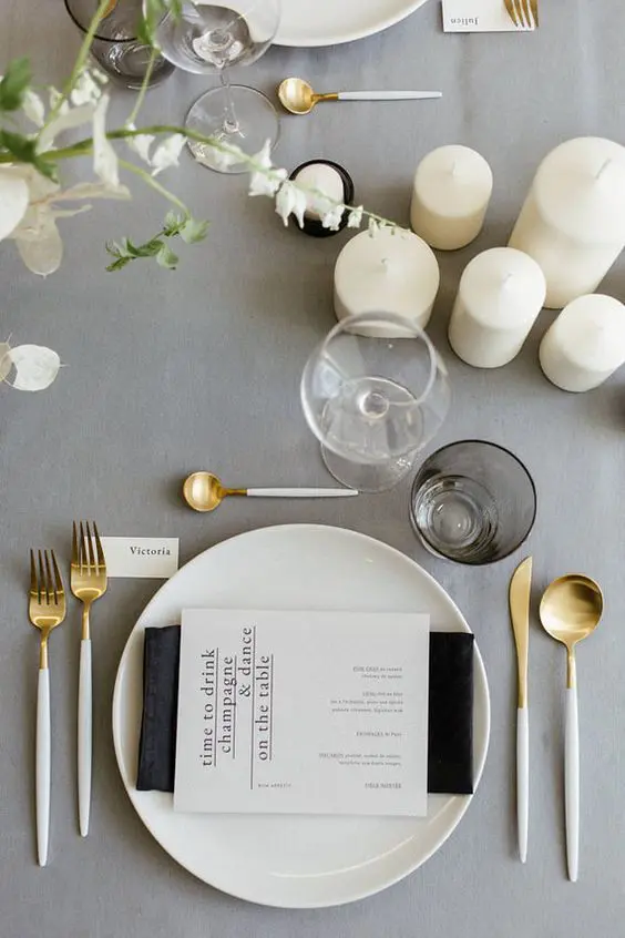 a beautiful minnimalist wedding tablescape with a grey tablecloth a black napkin white cutlery pillar candles and plates and menus