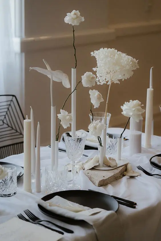 a bold and edgy minimalist wedding tablescape with neutral linens black chargers and cutlery neutral candles of various sizes and white blooms