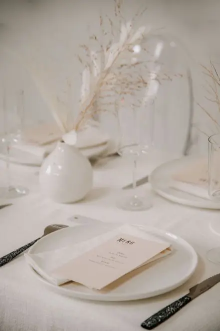 a chic minimalist and neutral wedding tablescape with dried grasses in white vases white plates and neutral menus and catchy cutlery