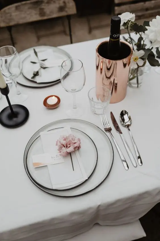 a chic minimalist wedding tablescape with elegant white linens white plates with black edges white blooms and candles