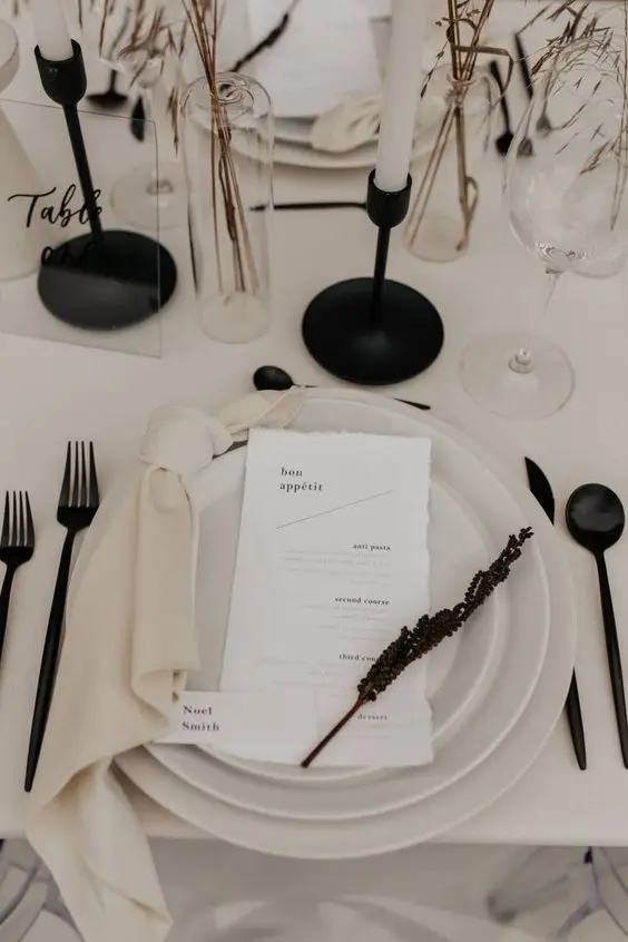 a contrasting minimalist wedding tablescape with neutral plates black cutlery candlesticks and dried blooms and grasses