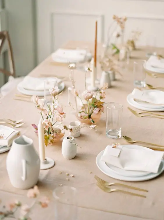 a delicate minimalist wedding tablescape with a blush tablecloth blush blooms and greenery neutral and rust candles white plates and napkins gold cutlery