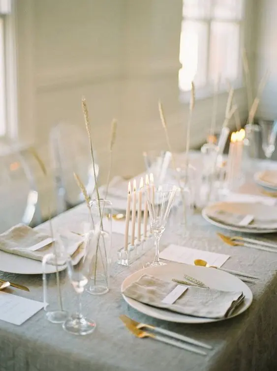 a delicate minimalist wedding tablescape with a grey tablecloth and napkins white chargers and cutlery some grasses in vases and tall and thin candles