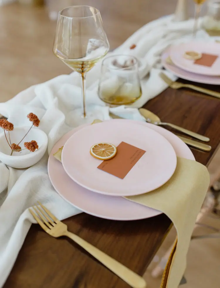 a minimalist bright wedding tablescape with a neutral runner pink plates mustard napkins and gold cutlery amber glasses