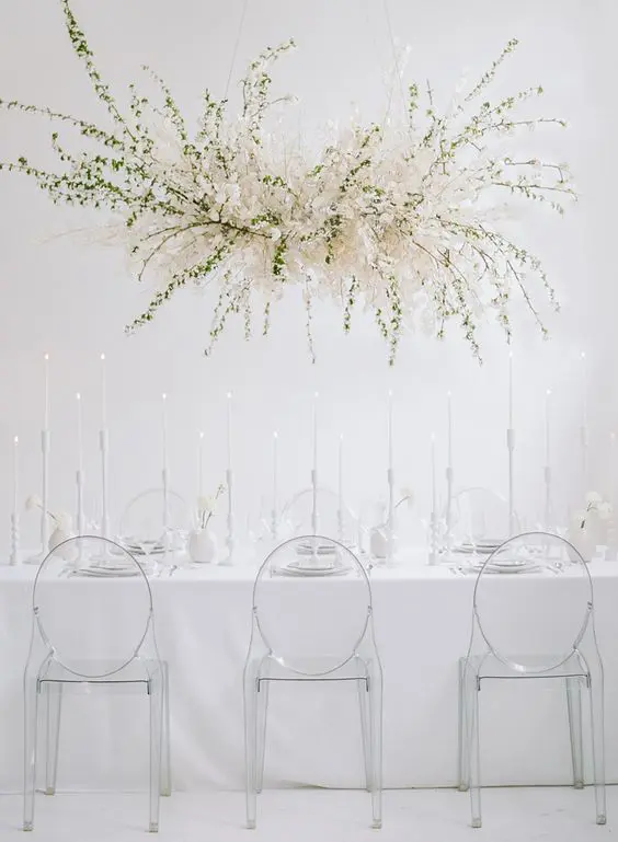 a minimalist ethereal wedding reception space with an oversized overhead floral decoration sheer chairs and everything white