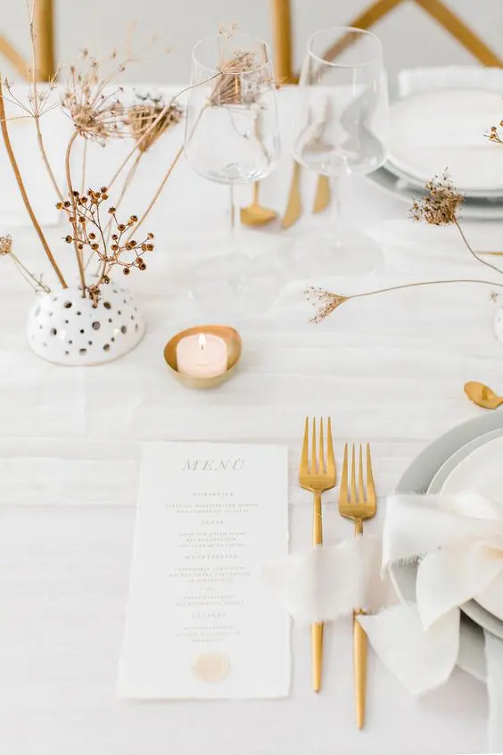 a minimalist neutral wedding table setting with white linens grey plates and chargers candles gold cutlery and dried blooms