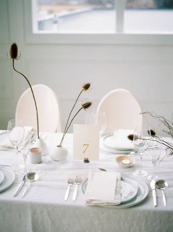 a minimalist neutral wedding tablescape with round white vases dried branches and blooms candles white plates and chargers and a linen tablecloth