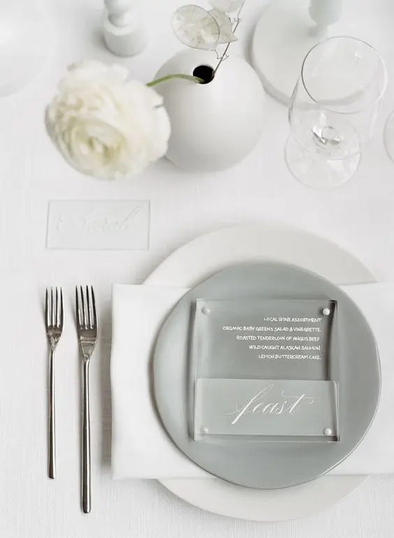 a neutral minimalist wedding table setting with white linens a grey plate white blooms in a round vase and simple cutlery