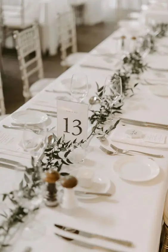 a neutral minimalist wedding tablescape with a greenery runner all crispy white around and white menus is a cool idea