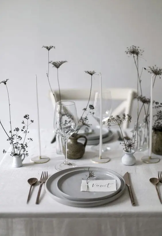 a neutral minimalist wedding tablescape with grey plates white vases with dried blooms simple cutlery and thin and tall candles