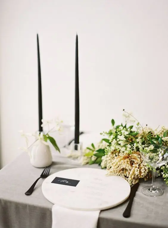 a pretty and elegant minimalist wedding tablescape with a grey tablecloth and white napkins a marble charger black candles and a greenery and white bloom centerpiece
