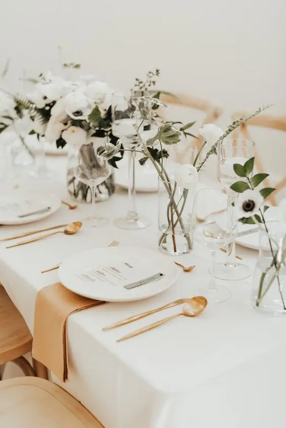 a pretty minimalist wedding tablescape with white blooms and greenery white plates gold cutlery and amber napkins plus floating candles