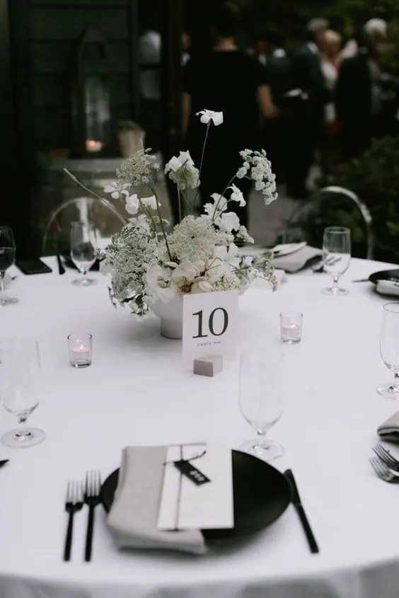 a sophisticated minimalist wedding tablescape with a crispy white tablecloth blakc plates and grey napkins neutral blooms and candles