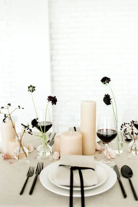 a stylish minimalist wedding table setting with grey linens white plates blush pillar candles black blooms and berries and black cutlery