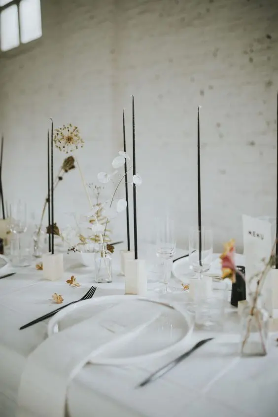 a stylish minimalist wedding tablescape with white plates and linens white candleholders tall and thin black candles and dried and fresh blooms
