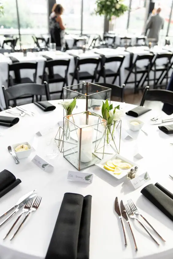 a stylish mnimalist wedding tablescape with candle lanterns with blooms and a candle with black napkins and a white tablecloth