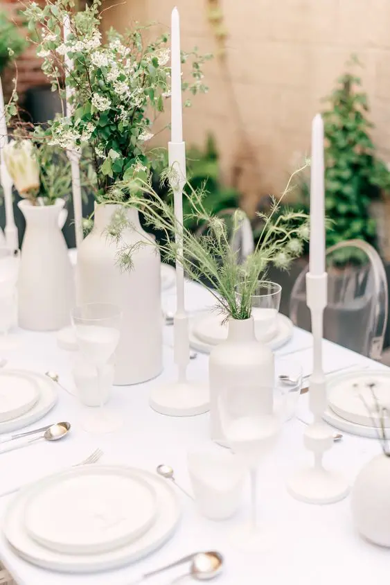 a very simple and minimal wedding tablescape with all white everything with greenery and white blooms tall and thin white candles and some cutlery