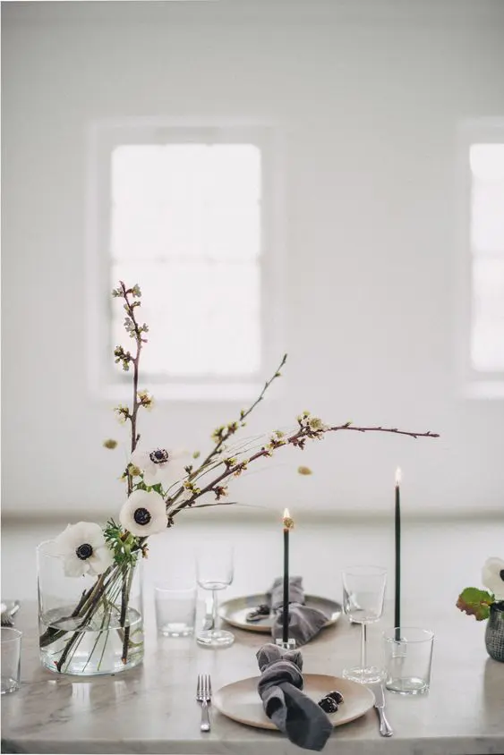 an elegant minimalist wedding table setting with neutral plates grey napkins white blooms thin and tall black candles