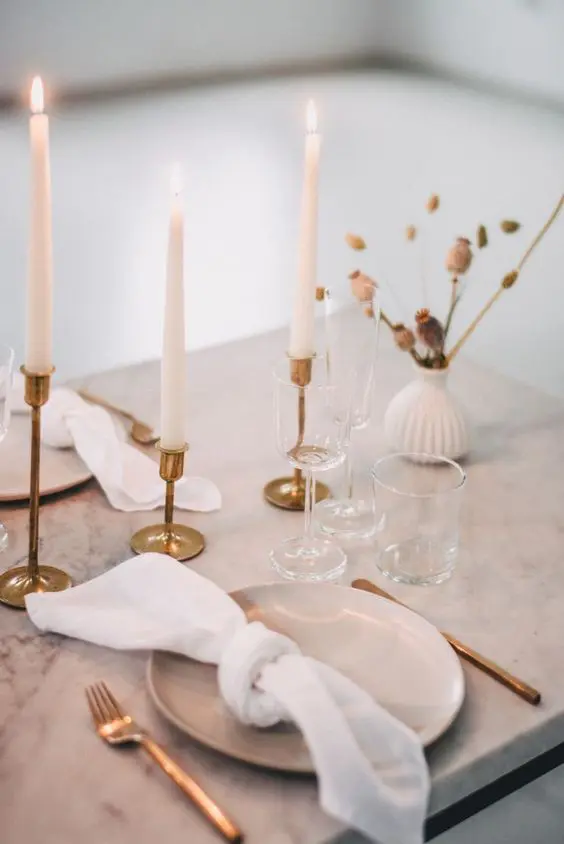 an elegant minimalist wedding tablescape with a marble table tall and thin candles dried blooms gold cutlery and gold candlesticks