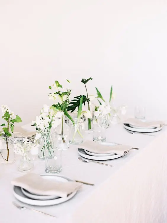 an ethereal minimalist wedding tablescape with all neutral everything and some white blooms and greenery in jars to form a centerpiece
