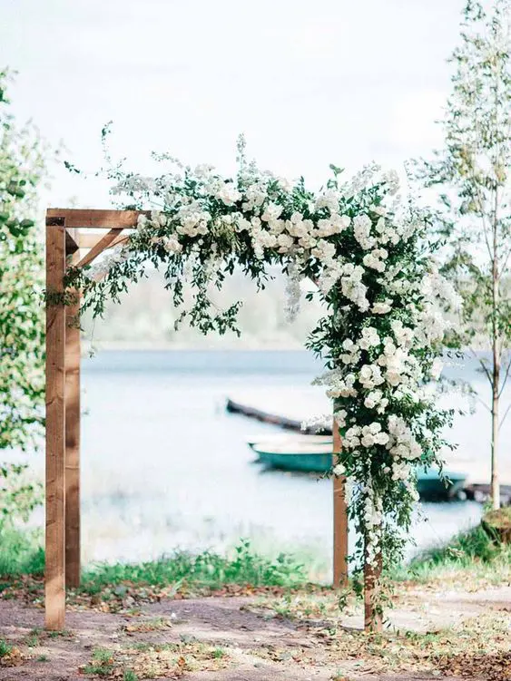 a chic rustic wedding arch of wood with greenery and white blooms on top is a very stylish and beautiful idea for any season
