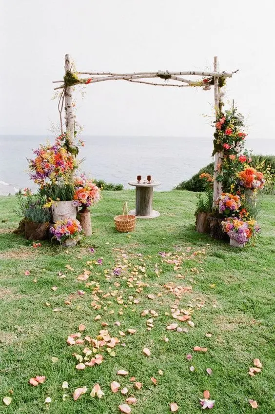 a colorful rustic wedding arch of birch branches greenery and colorful blooms petals on the ground and a sea view