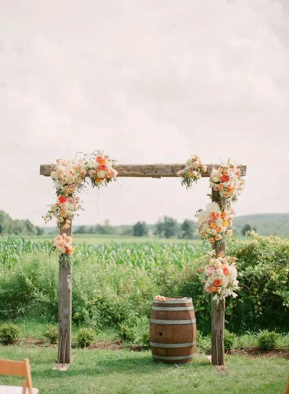 a colorful rustic wedding arch of rough wooden slabs and white and coral blooms plus a backdrop of a green field