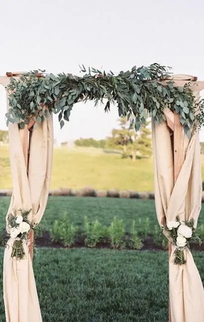 a cool rustic wedding arch covered with neutral burlapm eucalyptus white floral arrangements attached is a chic decoration