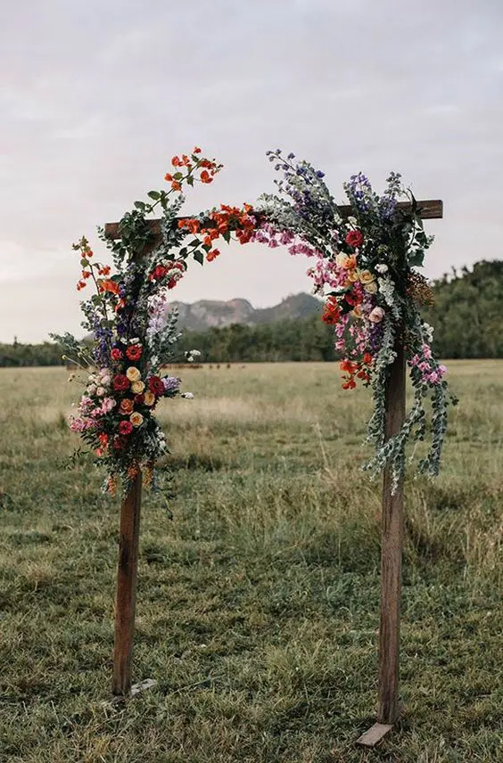 a cool rustic wedding arch of stained wood greenery and bright blooms is a lovely solution for a summer wedding
