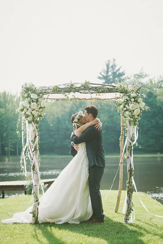 a pretty rustic wedding arch of brich branches with white ribbons white blooms and greenery and a cool backdrop of a pond
