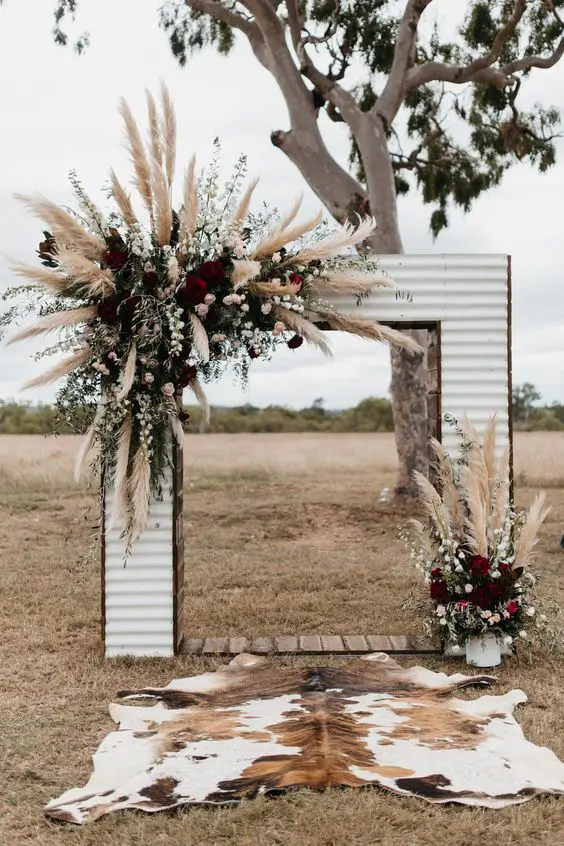 a rustic wedding arch covered with siding pampas grass white blush and dark purple blooms and greenery for a farmhouse wedding