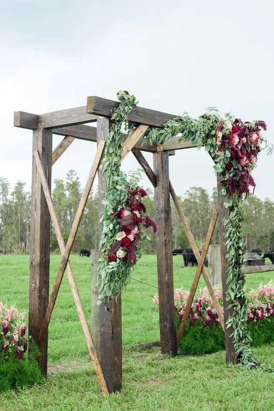 a rustic wedding arch decorated with greenery blush and burgundy blooms is a lovely idea for a fall rustic wedding