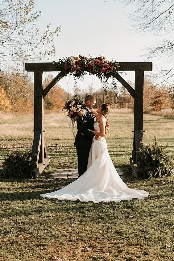 a rustic wedding arch decorated with greenery blush and dark blooms and dark greenery arrangements around