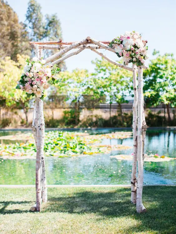 a rustic wedding arch of birch branches pastel blooms and greenery with a backdrop of a pond is a lvoely idea for summer