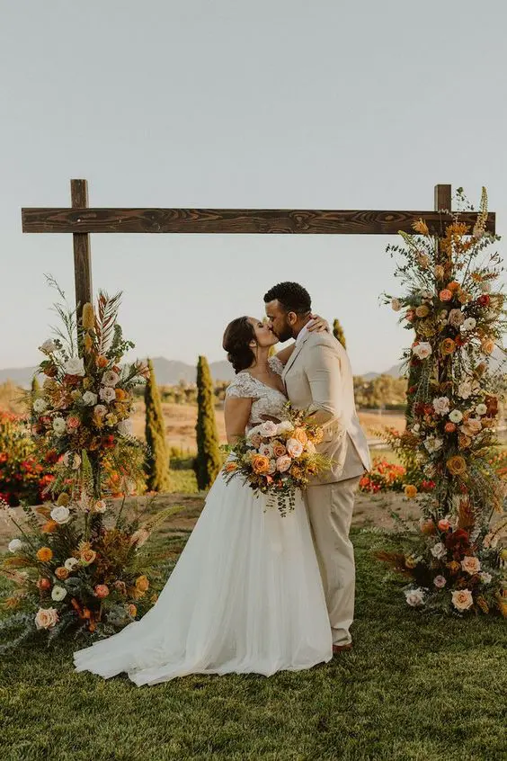 a rustic wedding arch of stained wood bright and blush blooms greenery pampas grass and twigs is very textural and dimensional