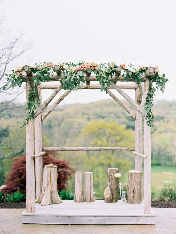 a rustic wedding arch with tree stumps greenery and peachy blooms plus a view of a fall forest for an autumn rustic wedding