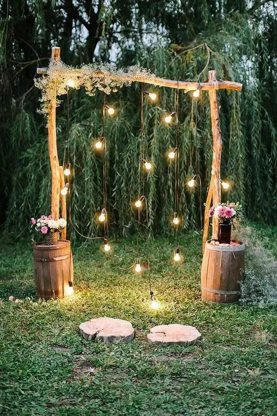 a simple rustic wedding arch of wooden branches some babys breath lights pink floral arrangements on barrels next to the arch