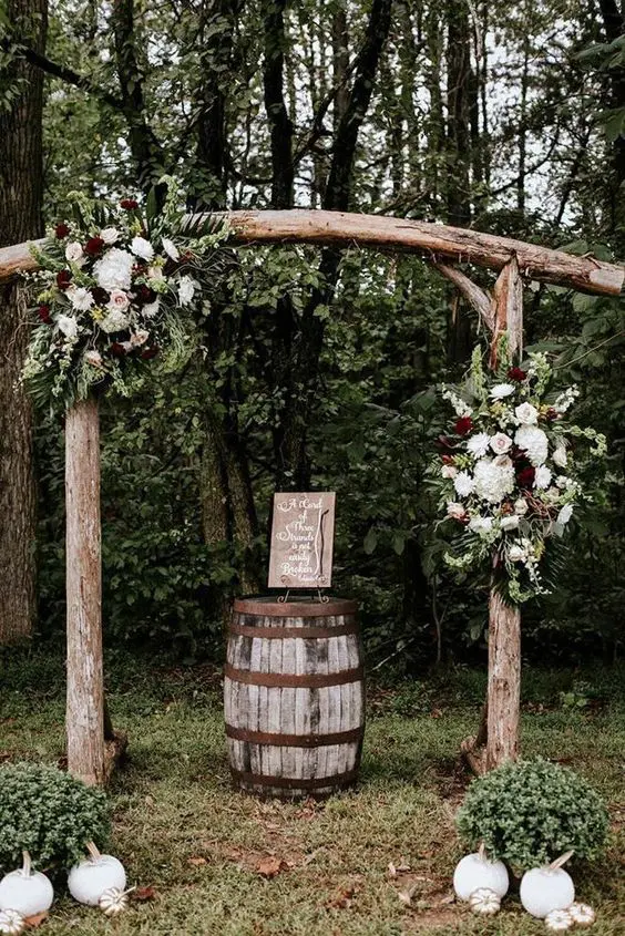 a simple rustic wedding arch with a curved top of logs white and burgundy blooms and greenery and white pumpkins on the ground