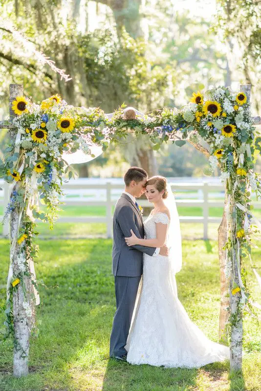 a summer rustic wedding arch of branches greenery sunflowers and ribbons plus some blue flowers is a very chic and cool idea