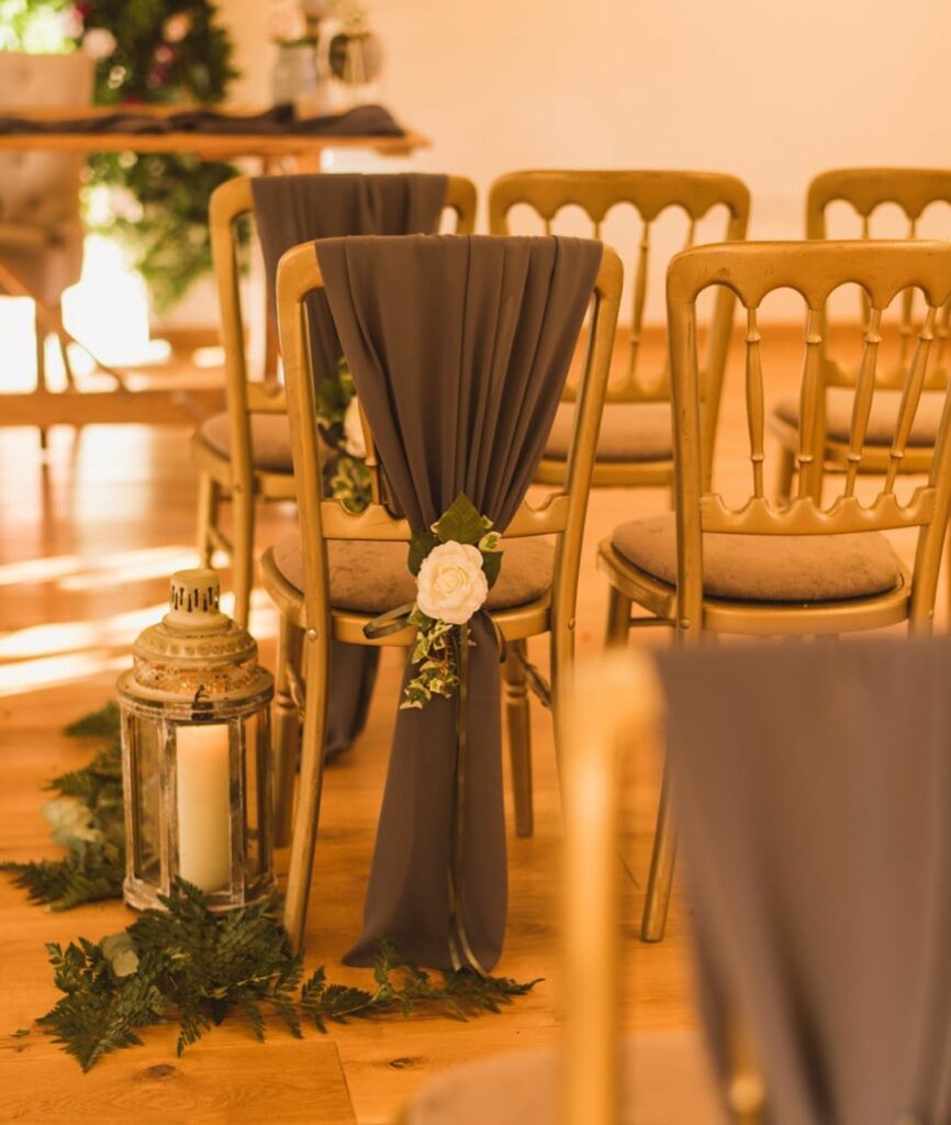 Here a rustic lantern illuminates a gorgeous aisle filled with a variety of plants. The aisle seats are wrapped in grey fabric decorated with ivory roses. Love the combination