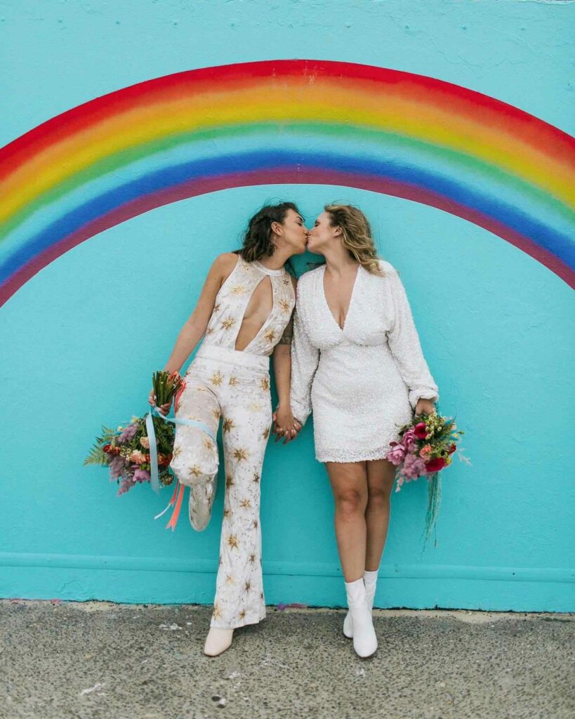 jumpsuit and short lesbian wedding outfits with rainbow backdrop and colorful bouquet