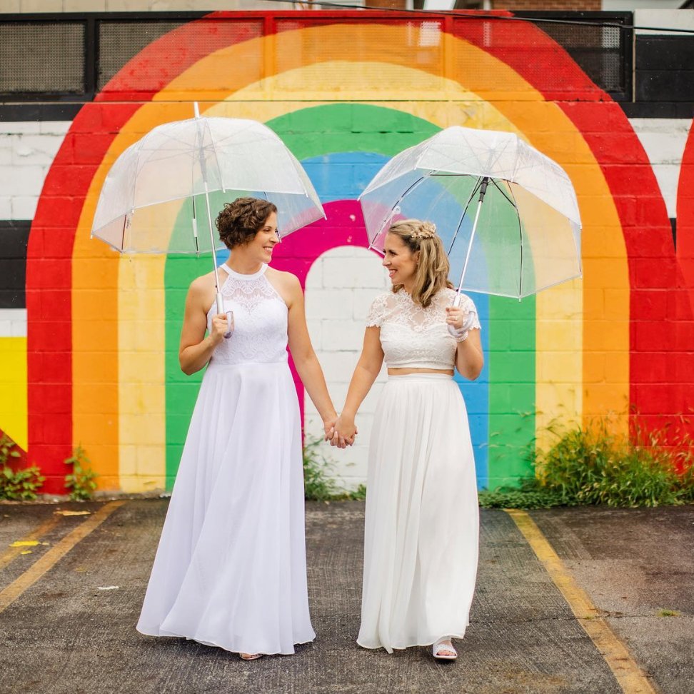 pride lesbian brides in gorgeous wedding dress with umbrella and rainbow backdrop